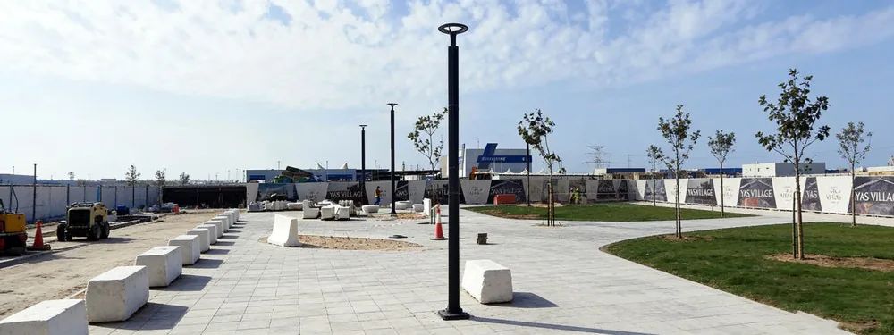 A sunlit outdoor plaza featuring modern black vertical pole lights, paved walkways, green lawns, young trees, and concrete blocks, with cons