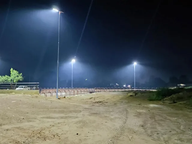A nighttime view of a dirt field illuminated by three tall streetlights against a dark sky.