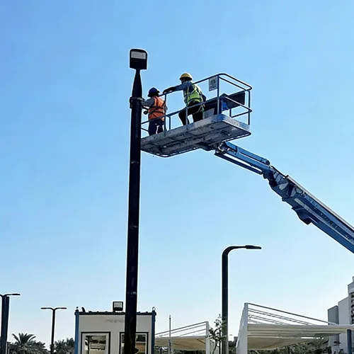 Two workers in safety gear on a lift inspect a tall street light pole against a clear blue sky.