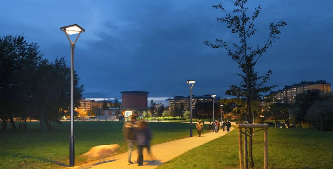 A park at dusk features a winding illuminated path with modern solar streetlights and pedestrians walking under a deep blue sky.