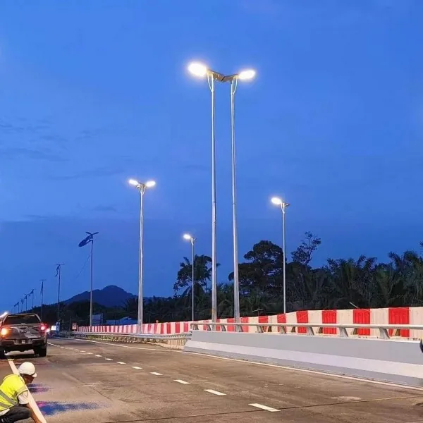 A newly constructed road at dusk is illuminated by tall double-headed streetlights, with a worker and pickup truck in the foreground.